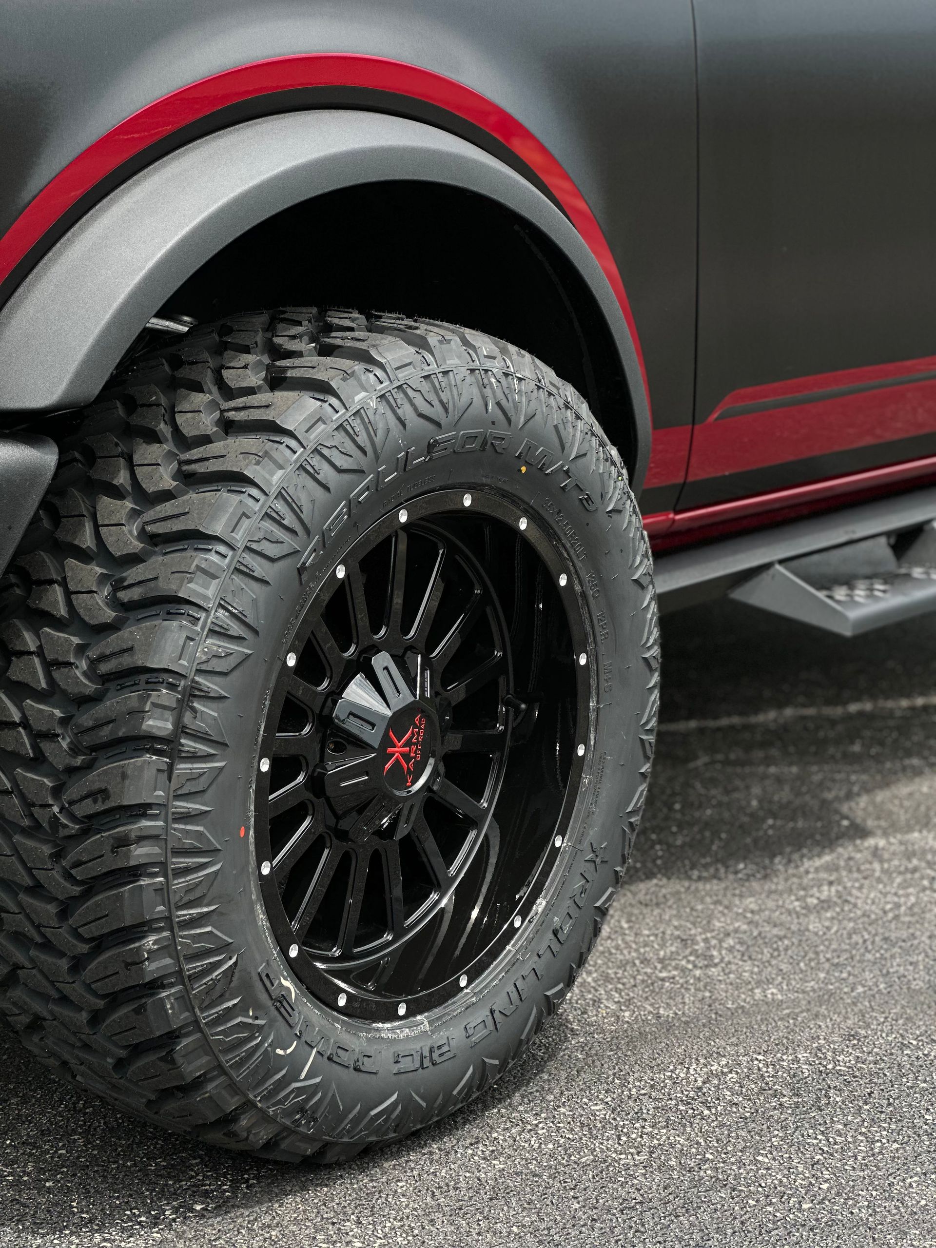 Close-up of a black off-road tire and wheel on a red vehicle, parked on pavement.