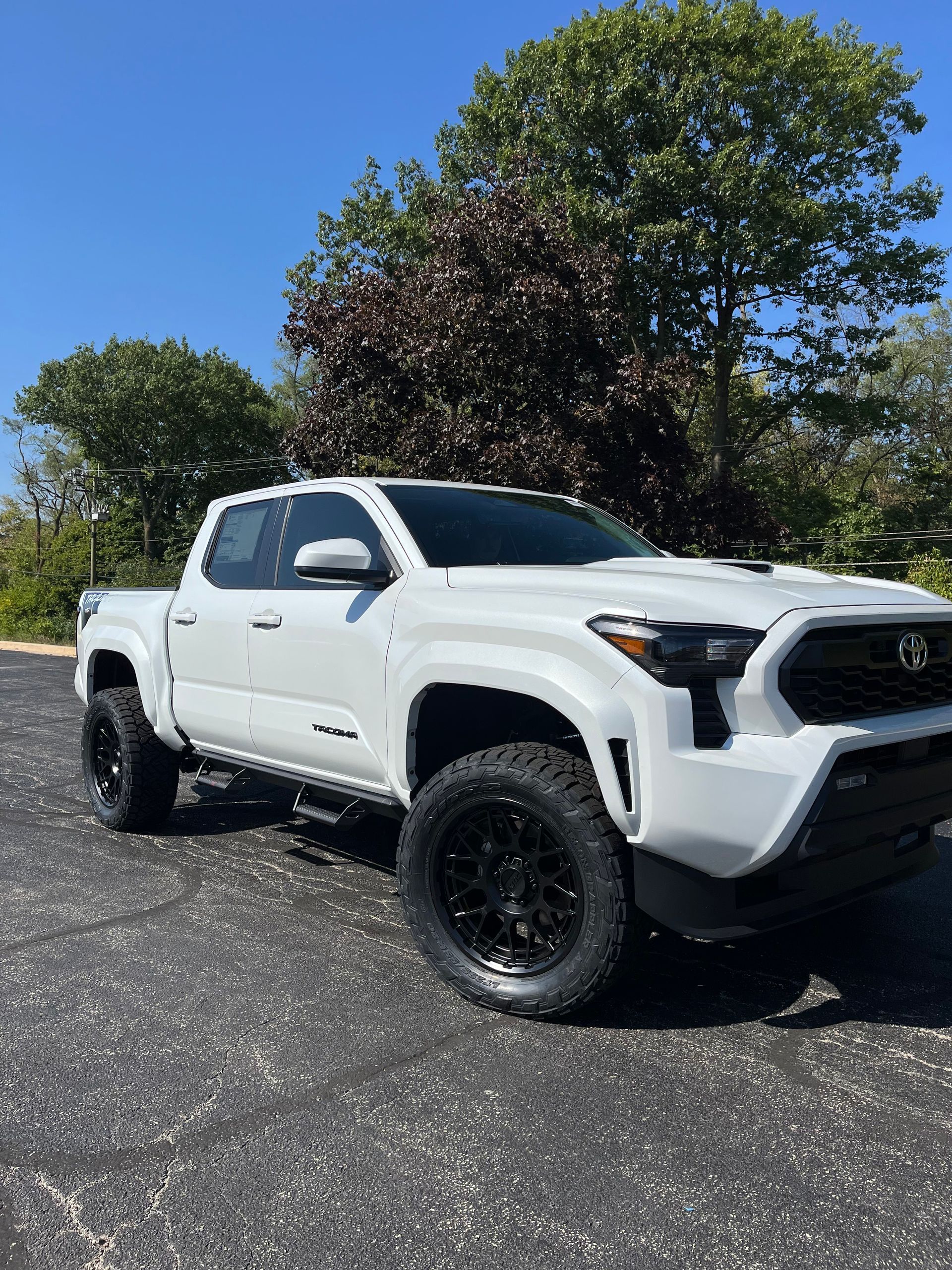 White lifted pickup truck with black wheels parked on a gravel lot beside trees under a clear blue sky