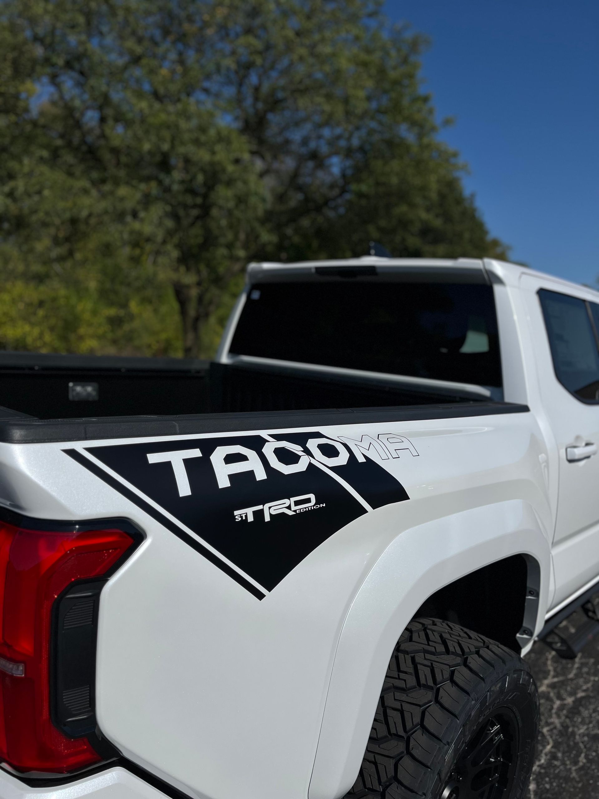White Toyota Tacoma pickup truck with black TRD badging, viewed from the rear side outdoors.