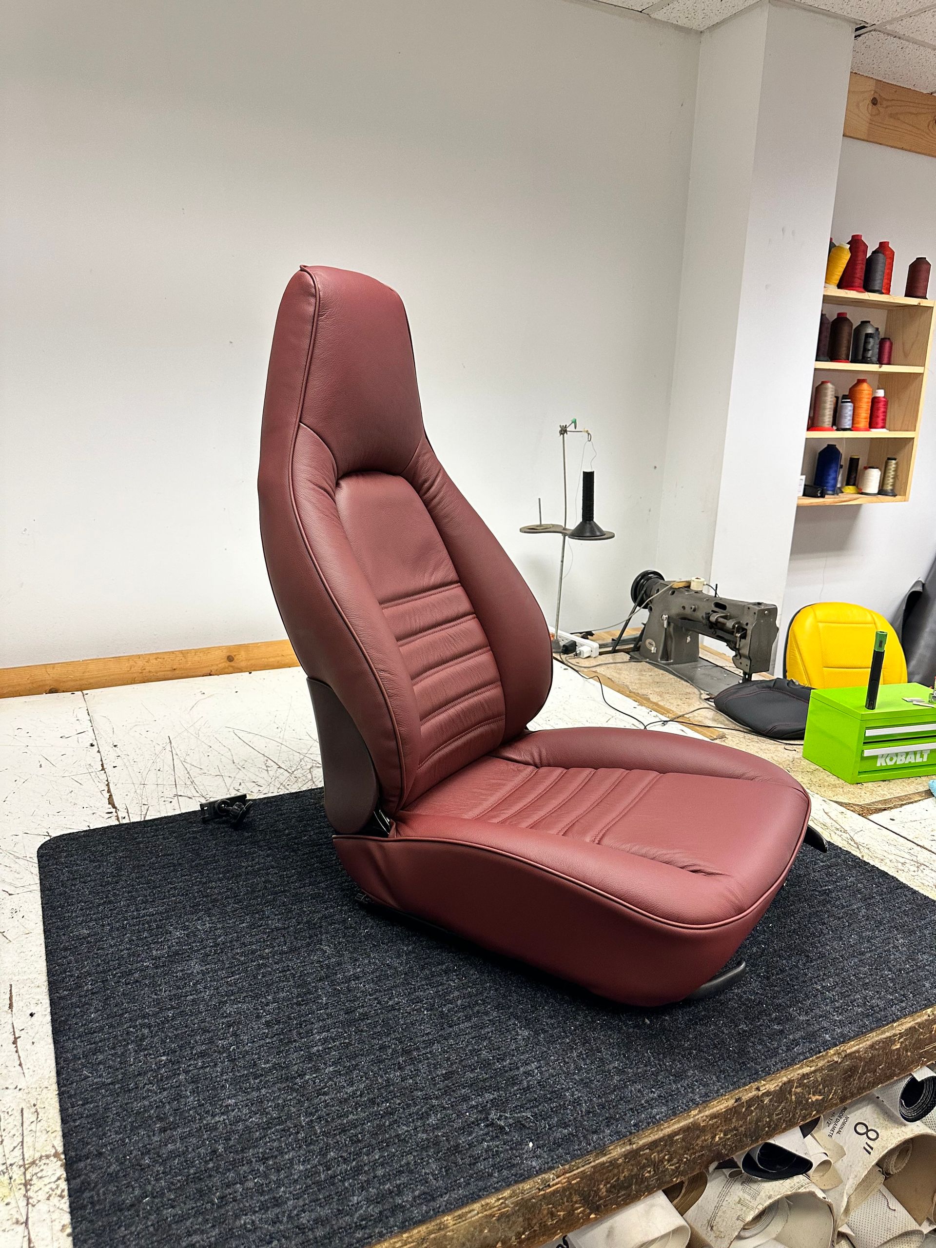 Maroon car seat mounted on a workbench in a workshop, with tools and supplies nearby.
