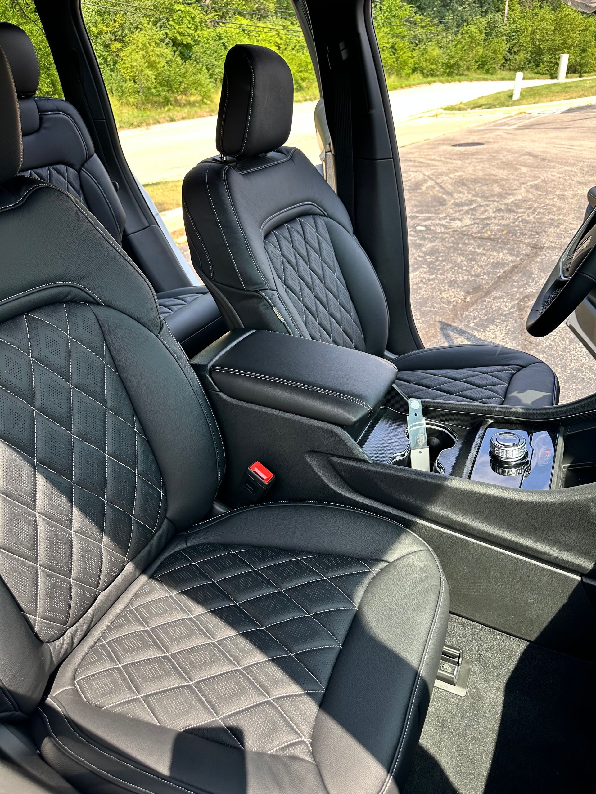 Black quilted car seats and center console inside a vehicle, with the passenger door open to a sunny road.
