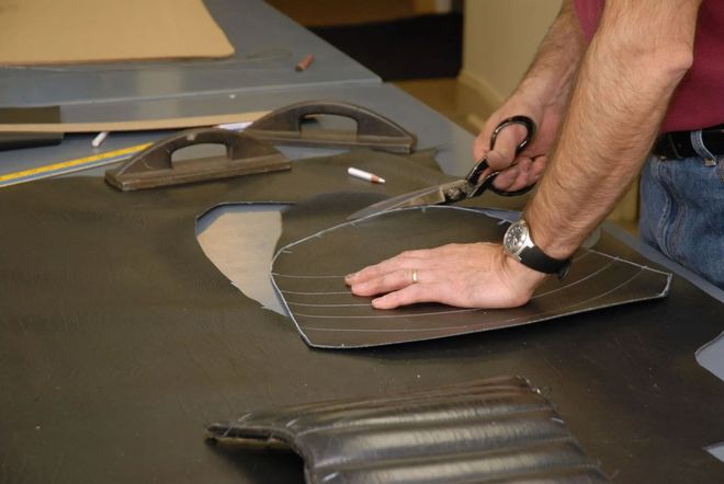 Person cutting black material with scissors on a worktable, with a leather car interior piece nearby