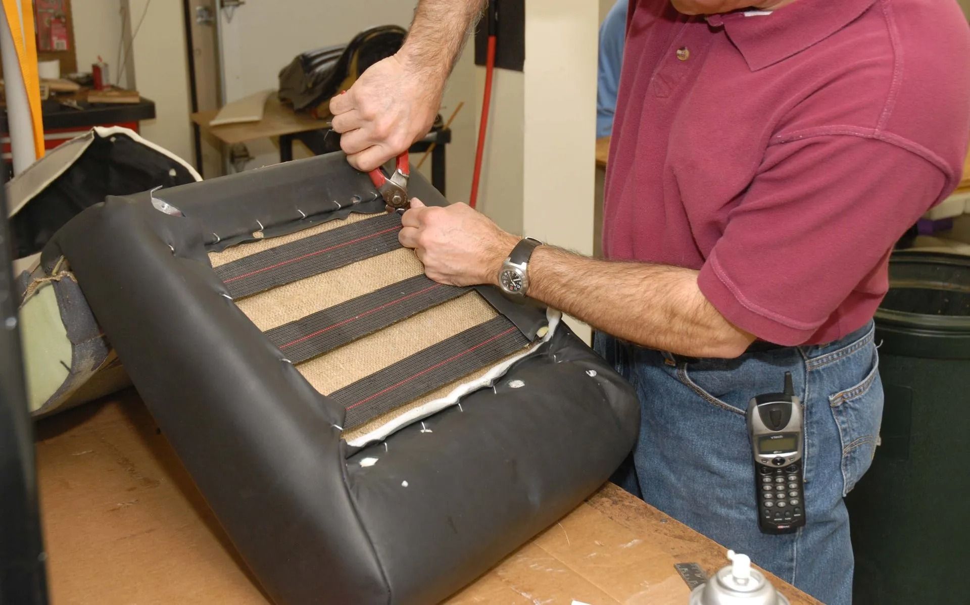 Man securing straps on a black chair cushion over a workbench, with tools nearby