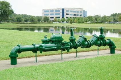 Green water pipes on display in a grassy area, with a lake and building in the background.