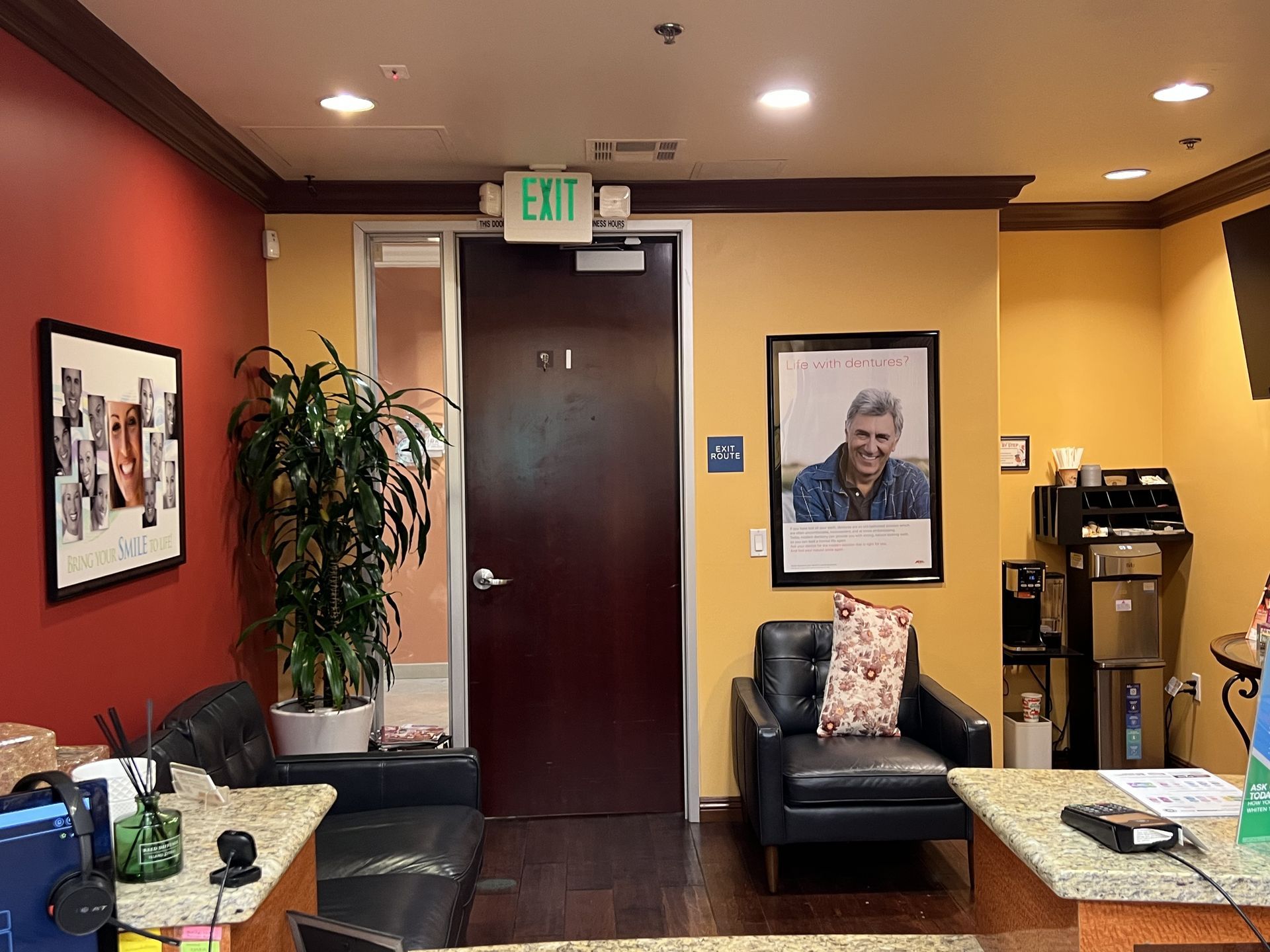Interior office waiting area, dark brown door with exit sign, framed art, leather chair, plants, coffee machine.