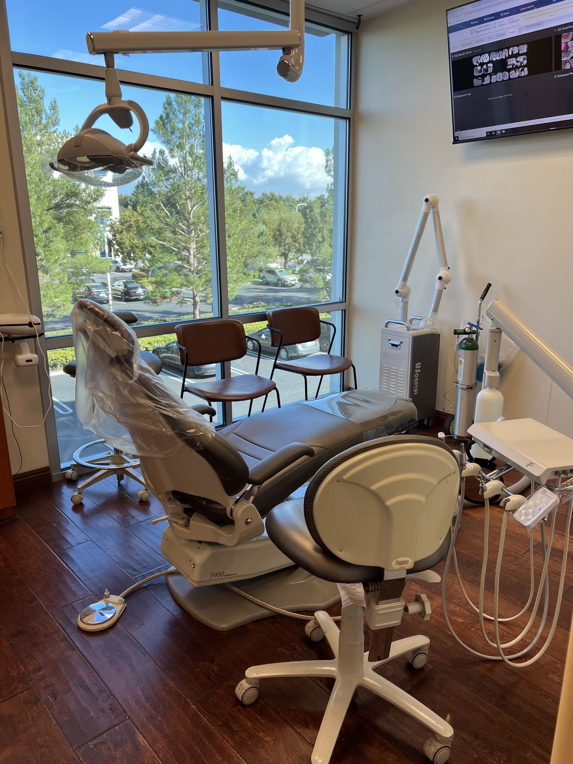 Dental examination room with chair, light, and equipment near a window overlooking trees.