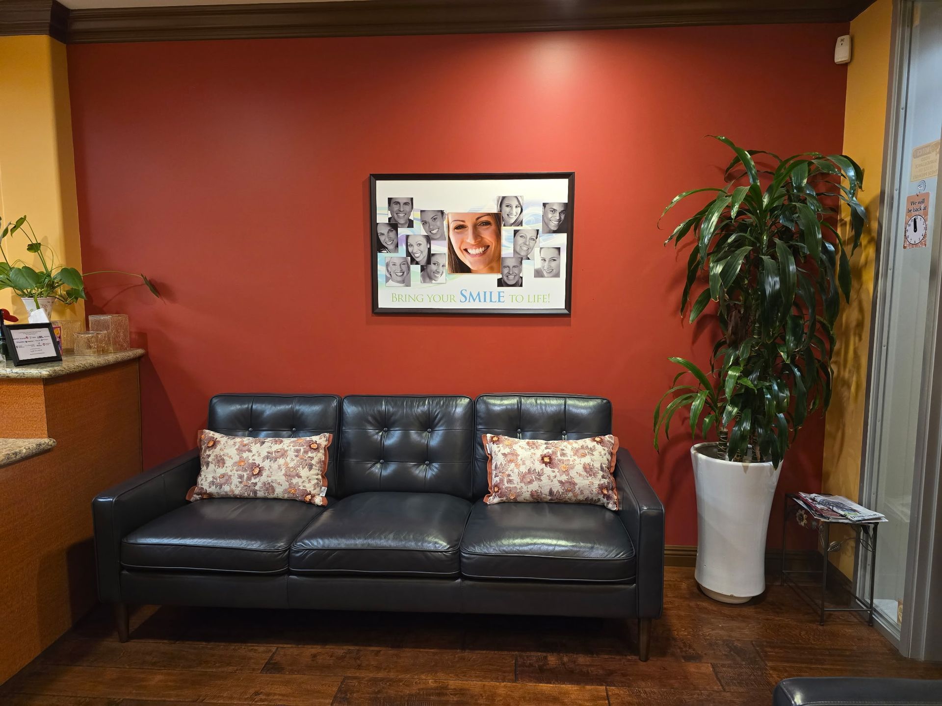 Dental office waiting room: black leather sofa, red wall, poster, potted plant.