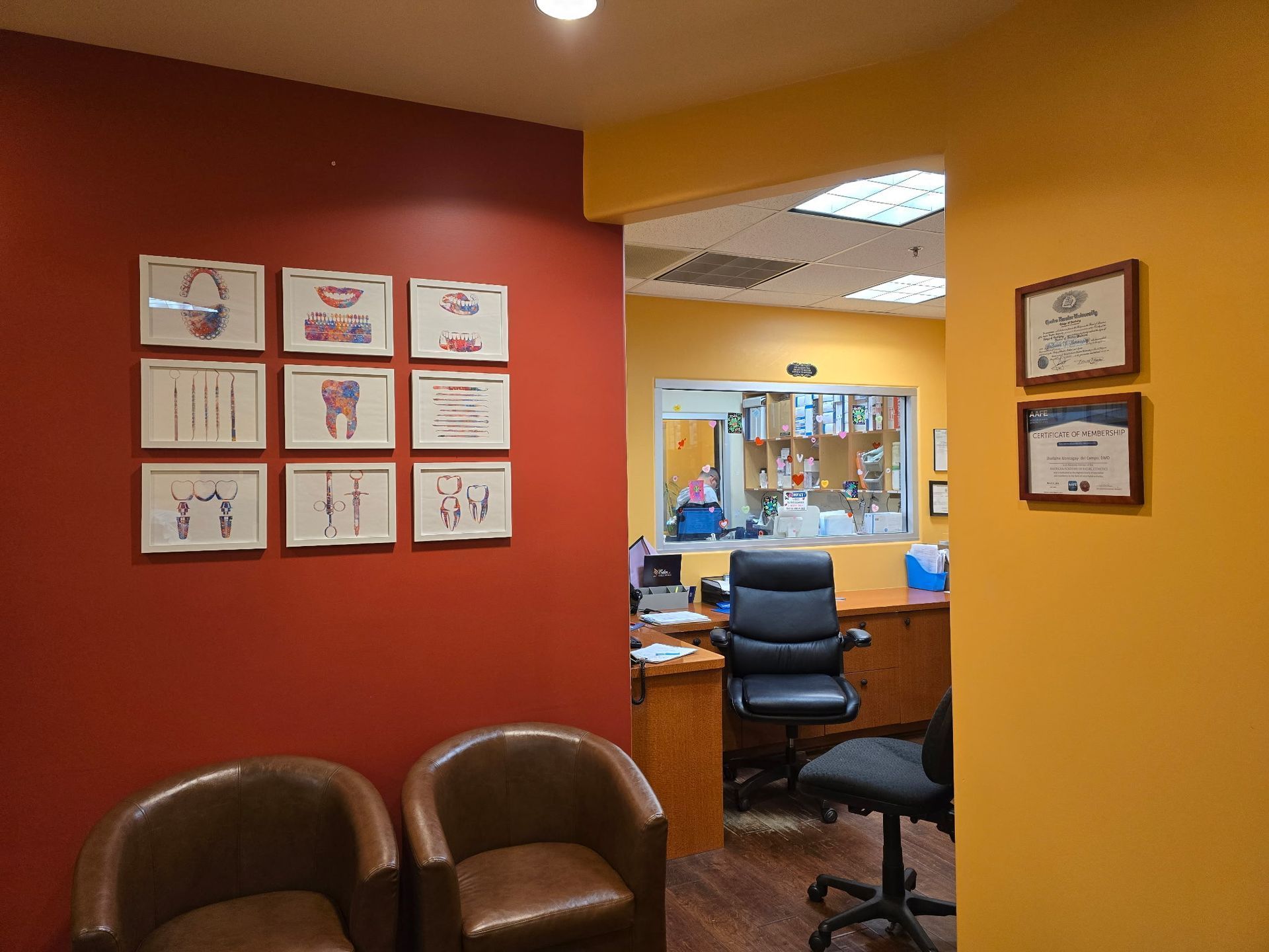 Waiting room with red and yellow walls, chairs, and diagrams. View into a bright office area.