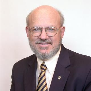 Portrait of a smiling man in a dark suit, striped tie, and glasses against a light background