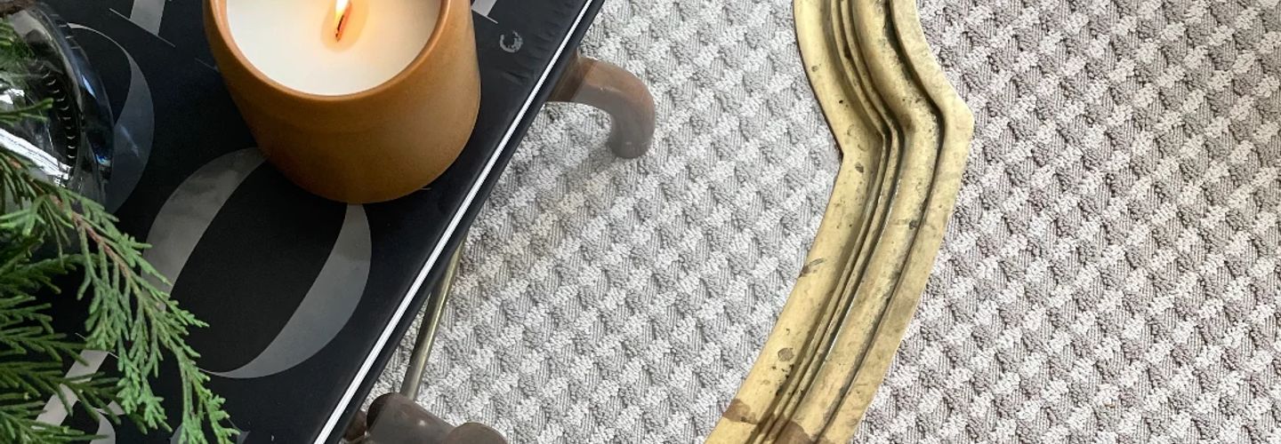 A candle in a brown holder sits on a black tray with a sprig of greenery; textured, light-colored rug.