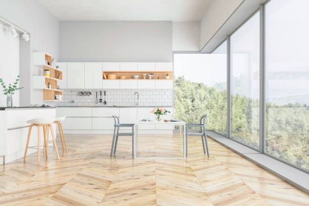 Modern white kitchen with large windows overlooking a forest, featuring a breakfast bar, table, and wooden floors.