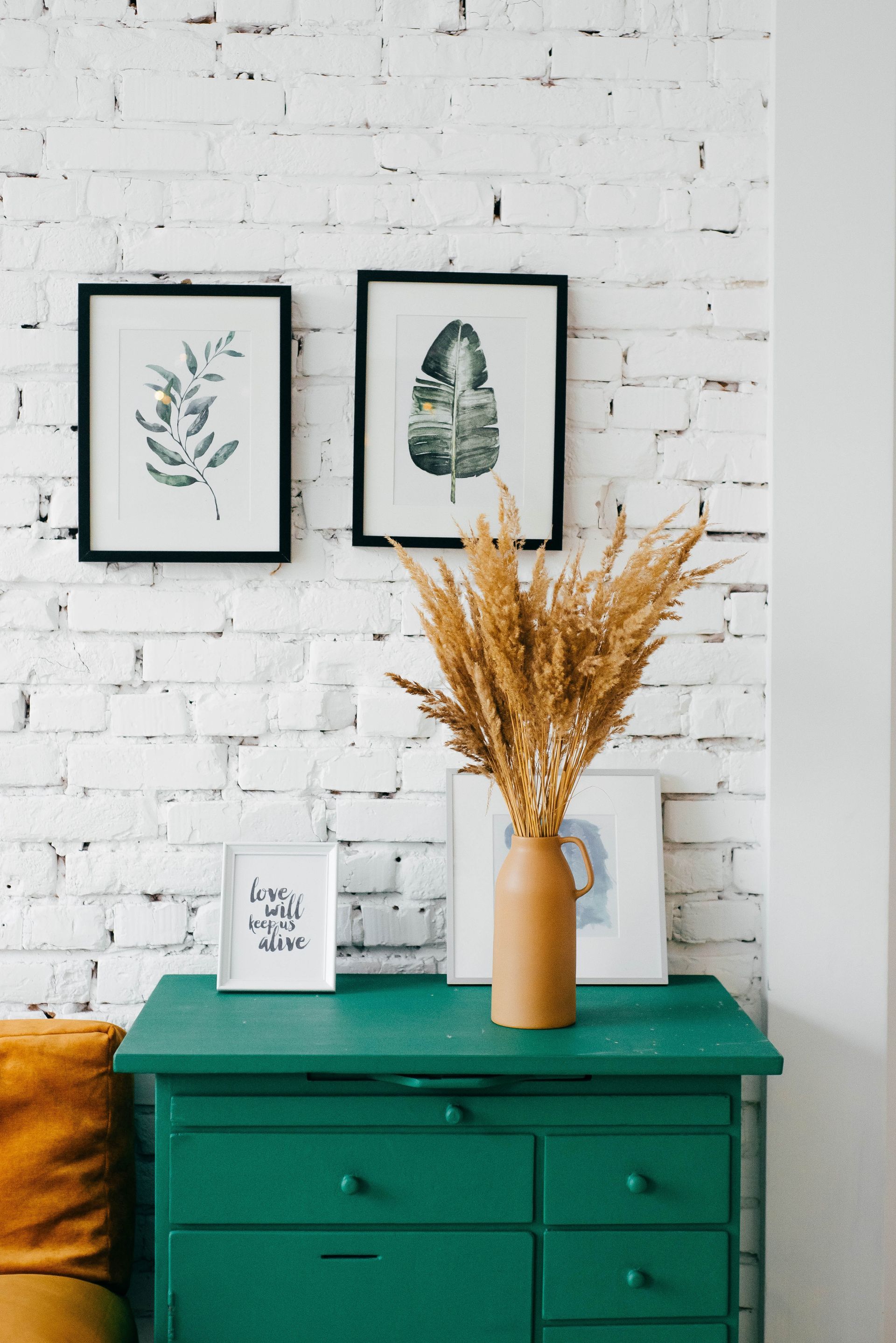 Green dresser with a vase of dried flowers, framed art on white brick wall.
