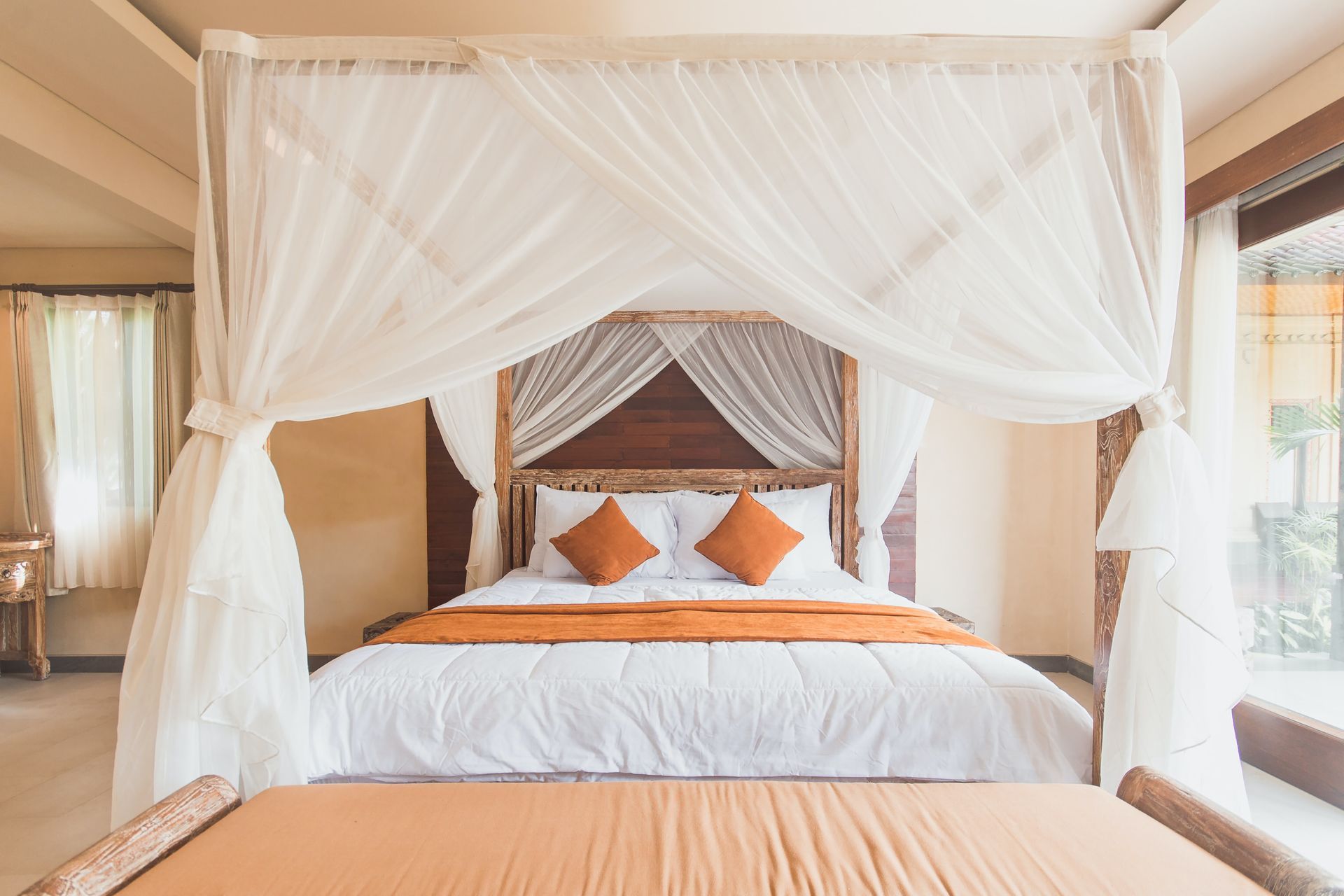 Bedroom with a four-poster bed draped in white fabric. Orange throw pillows and runner on the white bed, tan bench at the foot.