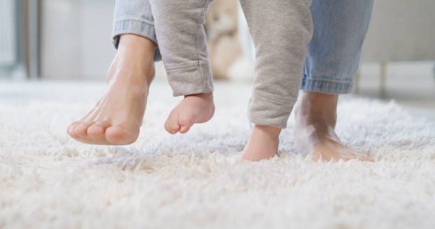 Adult supporting a toddler learning to walk on a fluffy white rug.