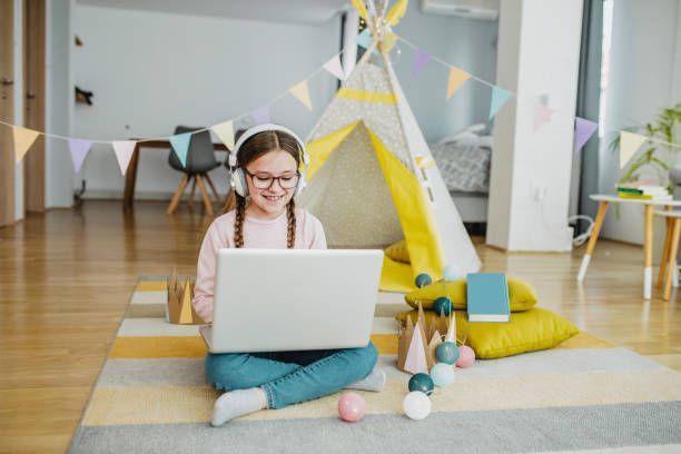 Girl with headphones uses a laptop while sitting on a rug, near a play tent.