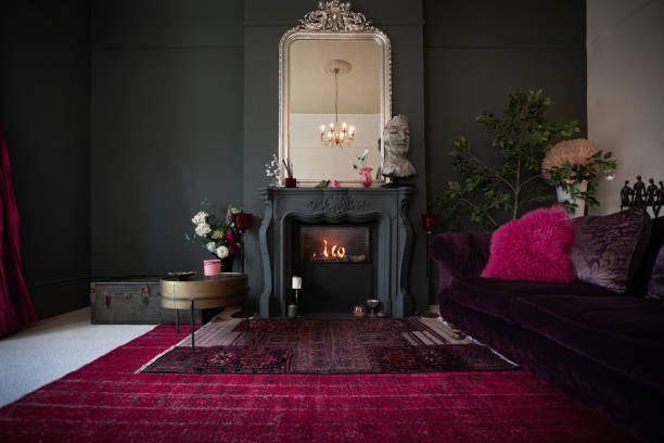 Dark-walled room with fireplace, ornate mirror, red rug and couch, flowers, and candles.