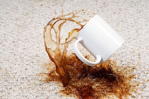 A white mug spills coffee onto a light-colored carpet, creating a brown stain.