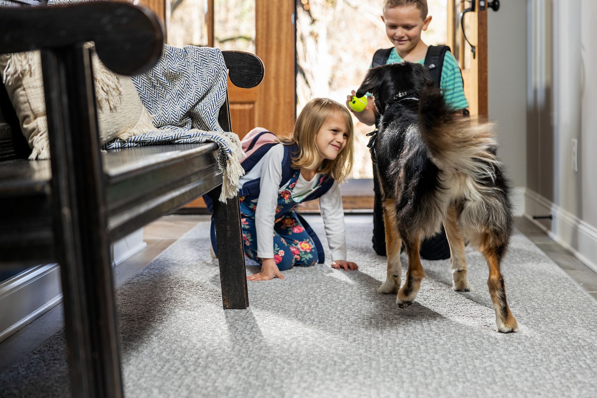 Two girls playing with a dog in a home entryway. One girl kneels, smiling; the other holds a ball.