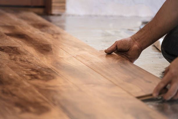 Hands installing wooden floor planks on a concrete surface.