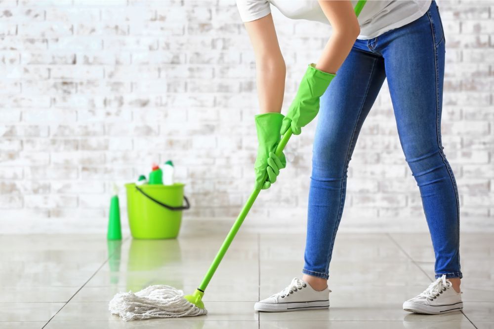 Person mopping a shiny, tiled floor wearing green gloves, white t-shirt, and jeans, with a bucket and cleaning supplies nearby.