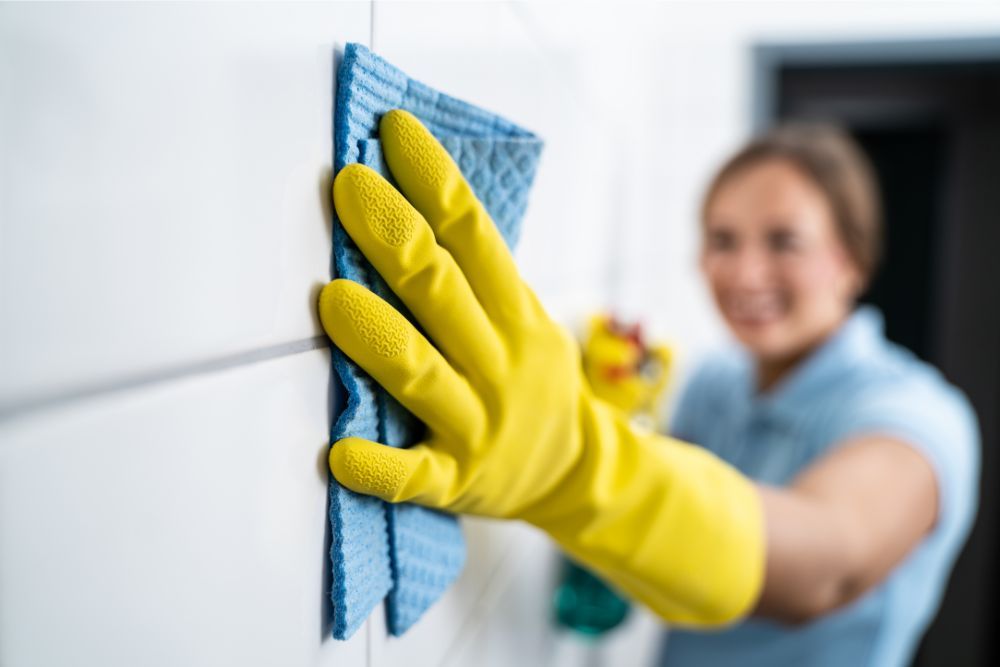 Person wearing yellow gloves wiping white tiles with a blue cloth.
