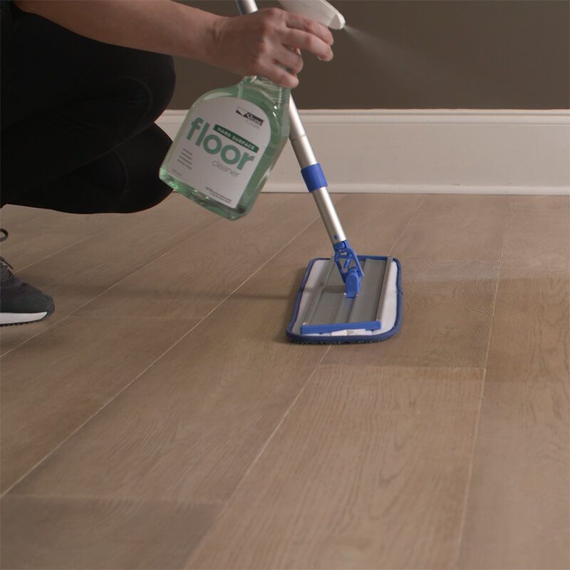 Person spraying cleaning solution onto floor, using a mop in a room.
