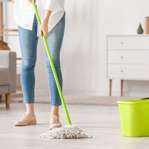 Woman mopping a light-colored wooden floor with a green mop and bucket in a living room.