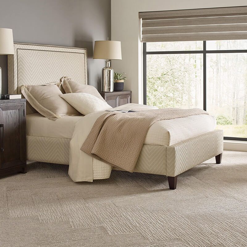 Bedroom with beige bed, nightstands, carpet, and large window with blinds.