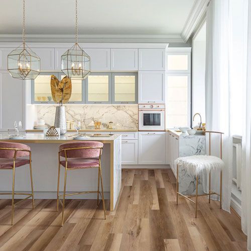 A modern kitchen with white cabinets, gold accents, pink stools, and marble countertops; wood floors.