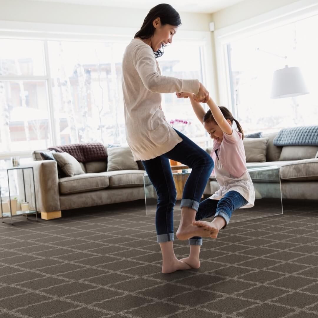 Woman and child playfully swinging, laughing in a living room with patterned carpet.