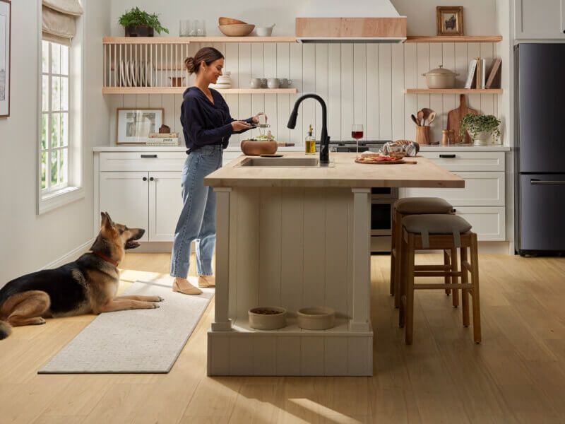 Woman in kitchen preparing food; dog watches. Kitchen has island, black faucet, and refrigerator.