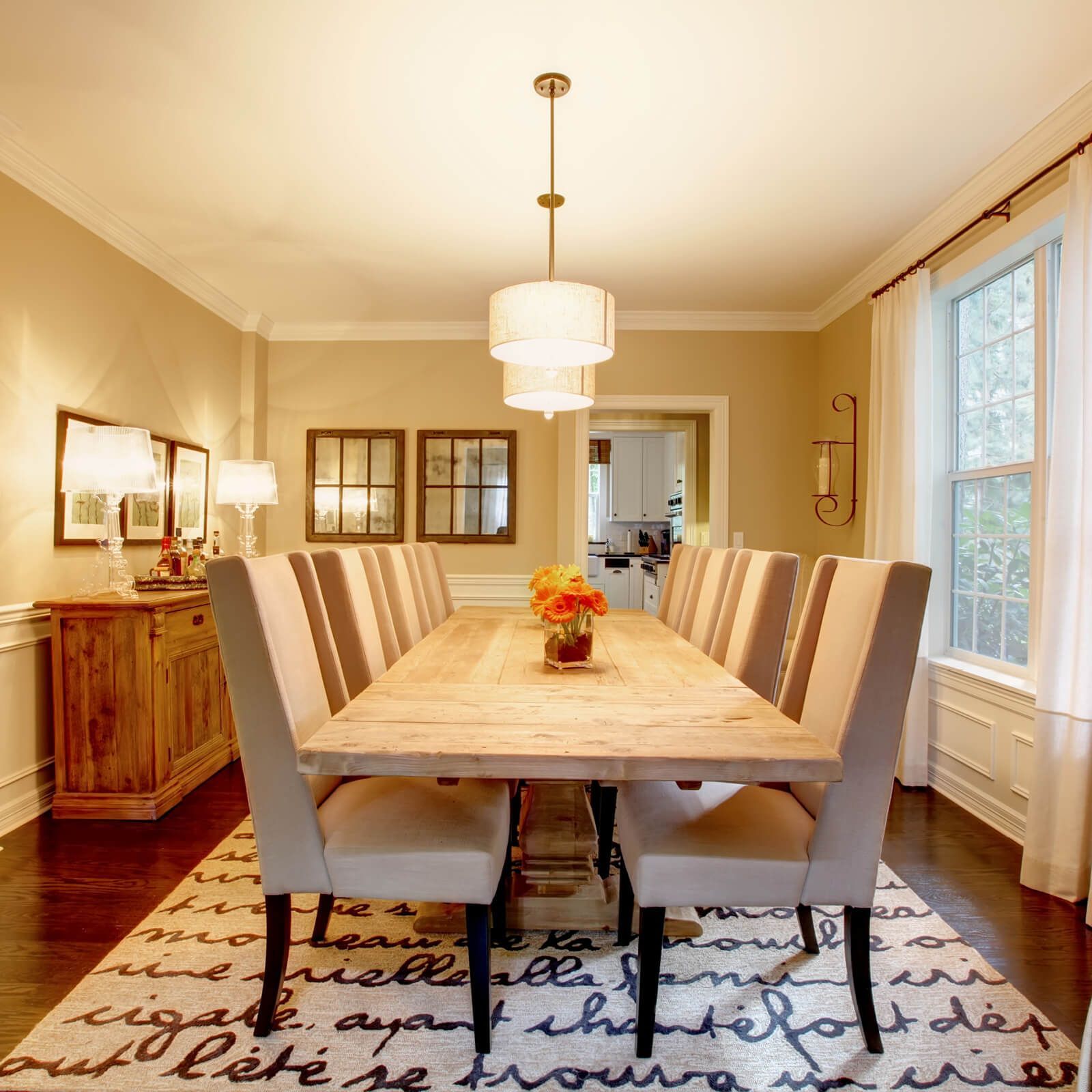 Dining room with long wooden table, chairs, and chandelier. Beige walls, window, and decorative rug.