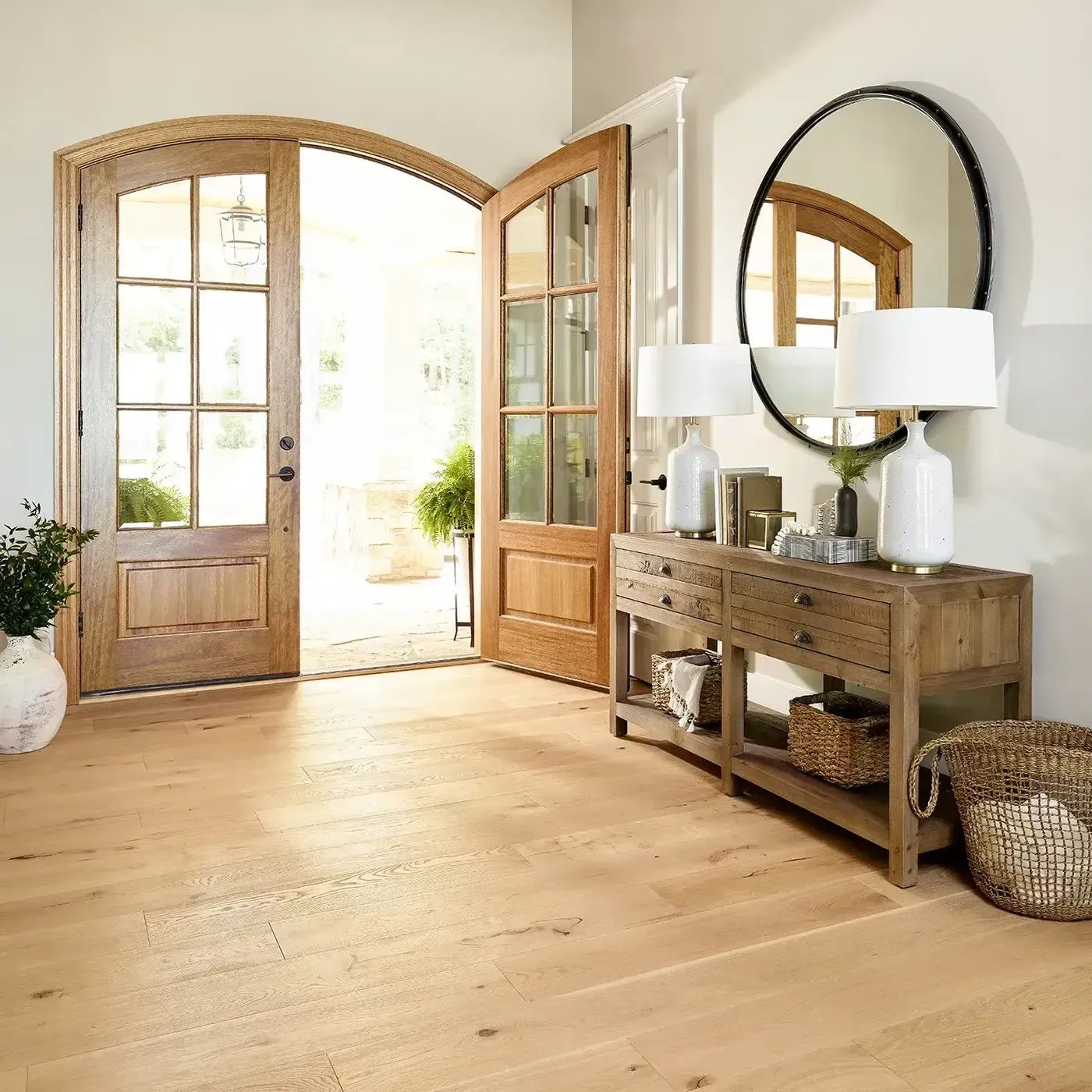 Wooden entryway with open double doors, wood console table, round mirror, lamps, and woven basket.