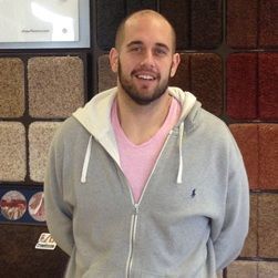 Man in gray hoodie and pink shirt smiles at the camera, standing in a carpet store.