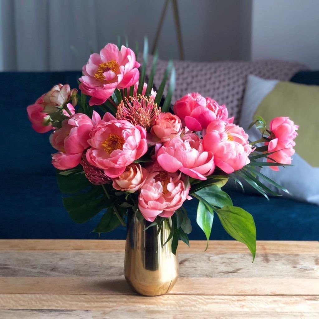 Pink peonies and greenery in a gold vase on a wooden table. A blue couch is in the background.