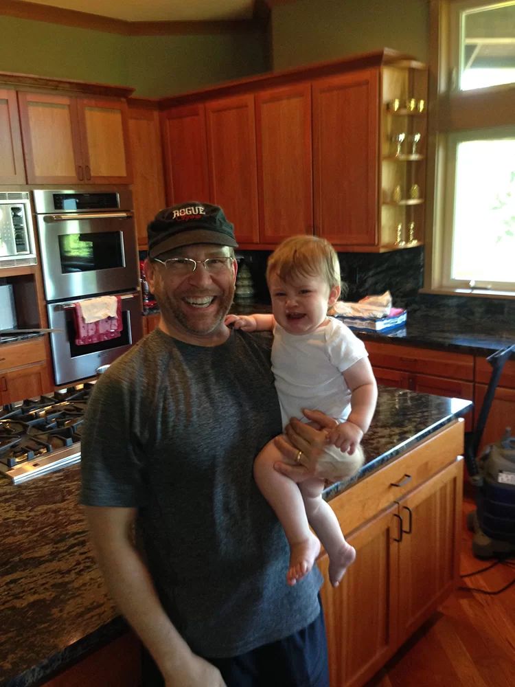 Smiling adult holding a laughing child in a warm kitchen with wooden cabinets and sunlight.