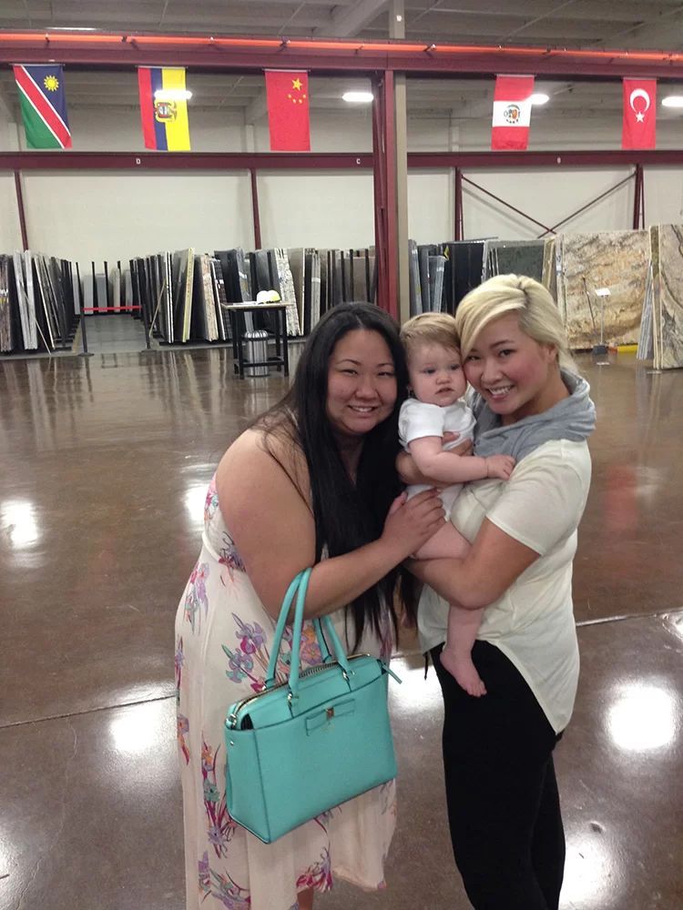 Three people smiling and holding a baby in a spacious indoor hall with flags overhead.