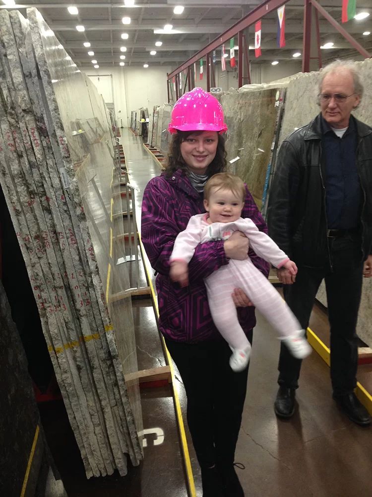 Woman in pink hard hat holding a baby in a warehouse aisle, with an older man beside her.