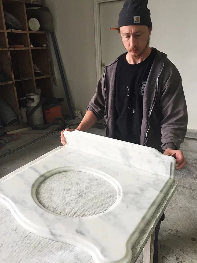 Person in a workshop holding a white carved stone countertop with a circular sink cutout.