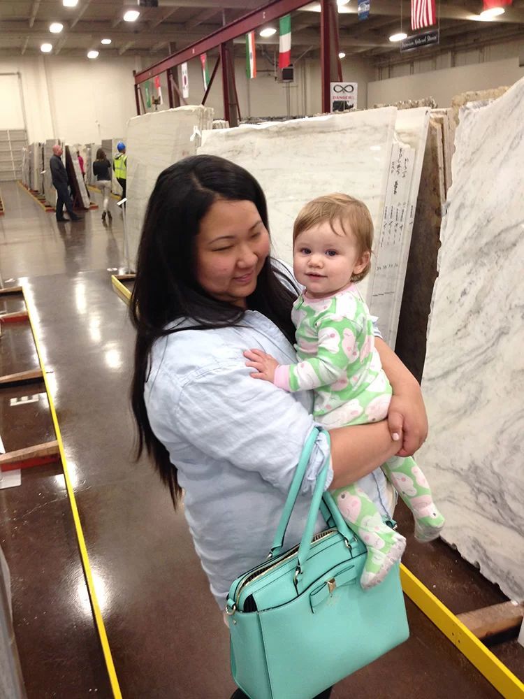 Woman holding a smiling toddler in a warehouse aisle, with large rolled materials stacked along the sides.