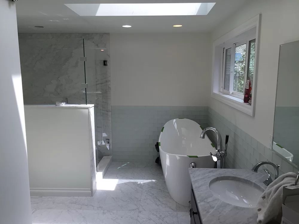 Bright bathroom with tub, sink, shower, and tiled walls, lit by a skylight and window.