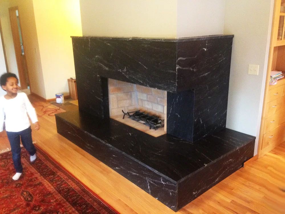 Modern black marble fireplace in a hardwood room, with a red rug and a child standing at left.