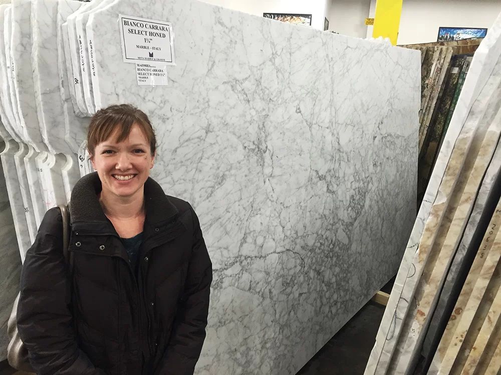 Smiling woman standing beside a large gray marble slab in a stone warehouse