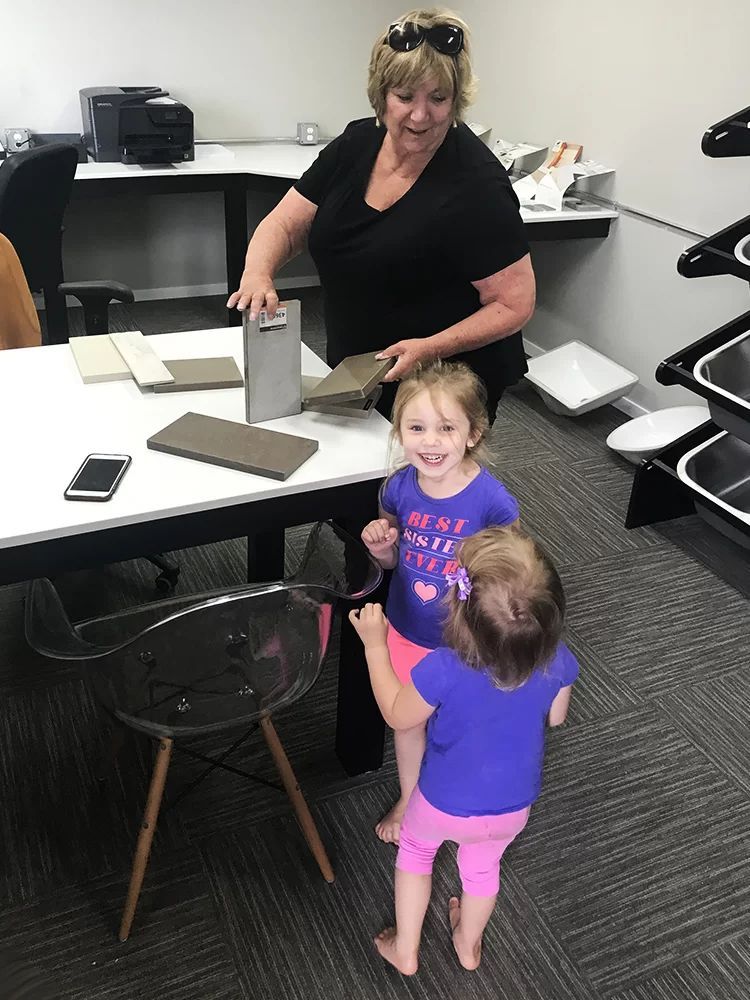 Two children stand by a desk while a smiling woman reaches toward items in an office.