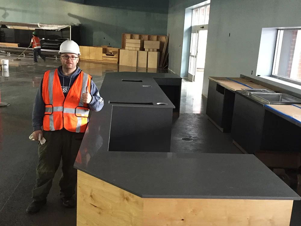 Worker in hard hat and orange vest standing in a workshop beside large black countertops.
