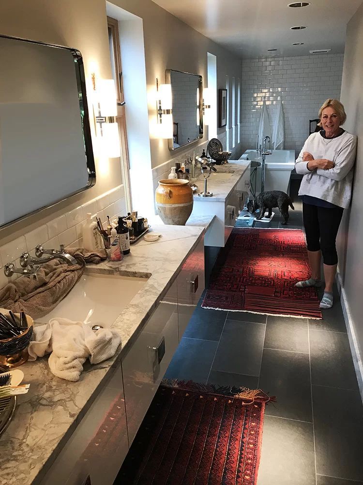 Long bathroom with double sinks, lit mirrors, and a woman standing near the shower at the far end.