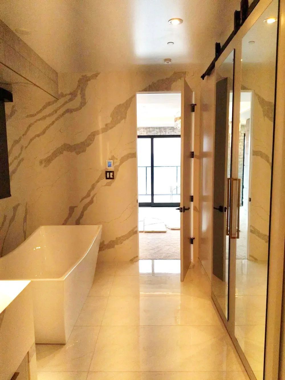 Modern bathroom with a bathtub, glass shower, and marble-tiled walls leading to a bright window.