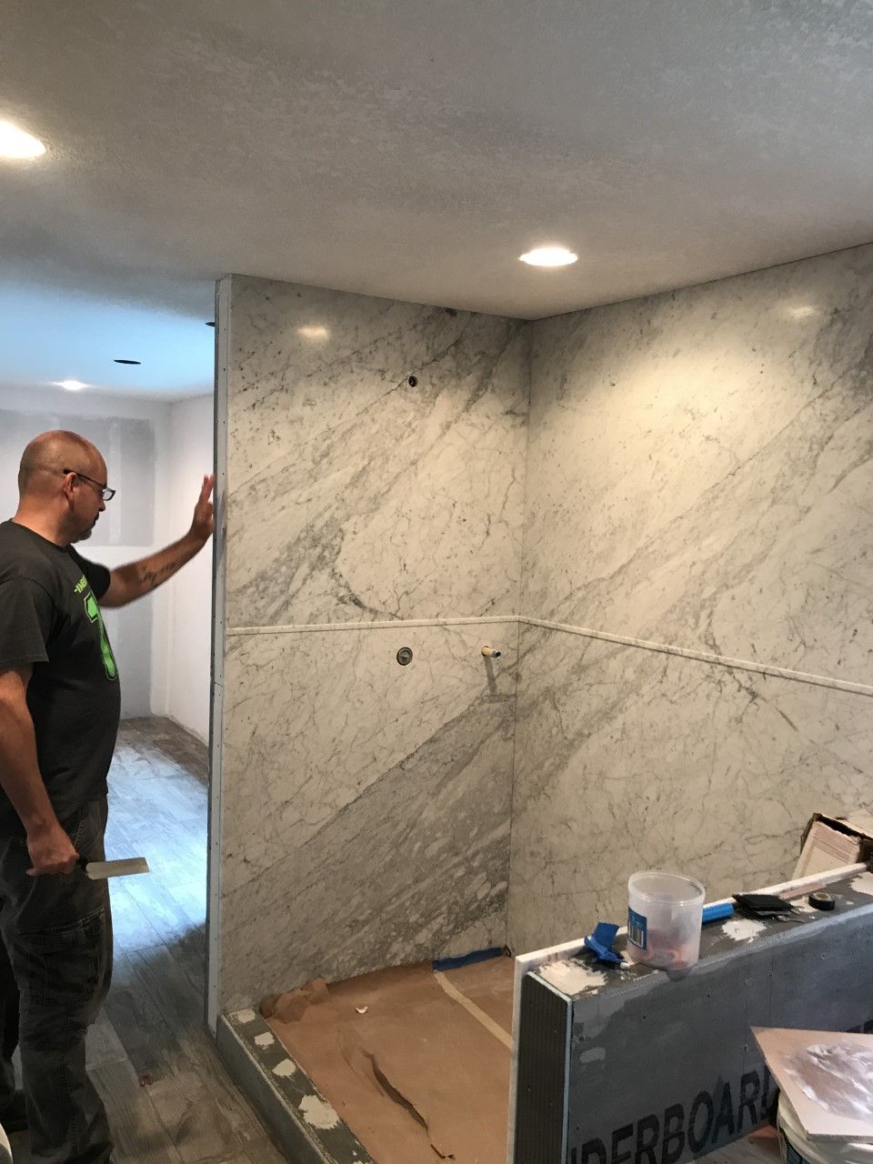 Man inspecting a marble-tiled wall in an unfinished room with exposed flooring and tools nearby