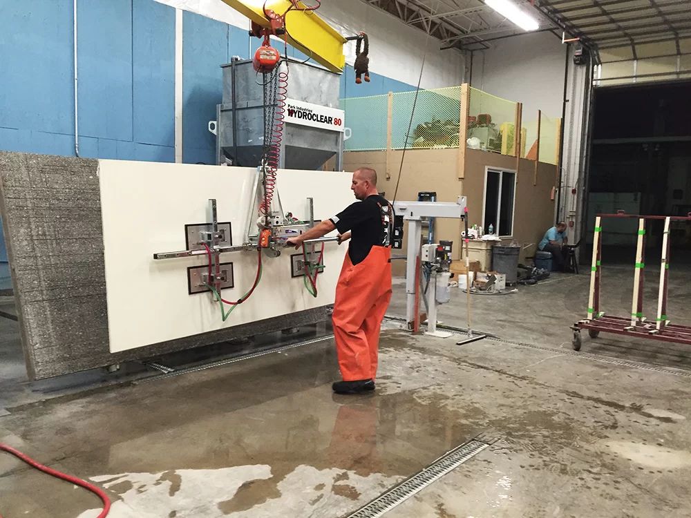 Worker operating a wall-mounted industrial lift in a wet workshop, with orange pants and a yellow crane overhead