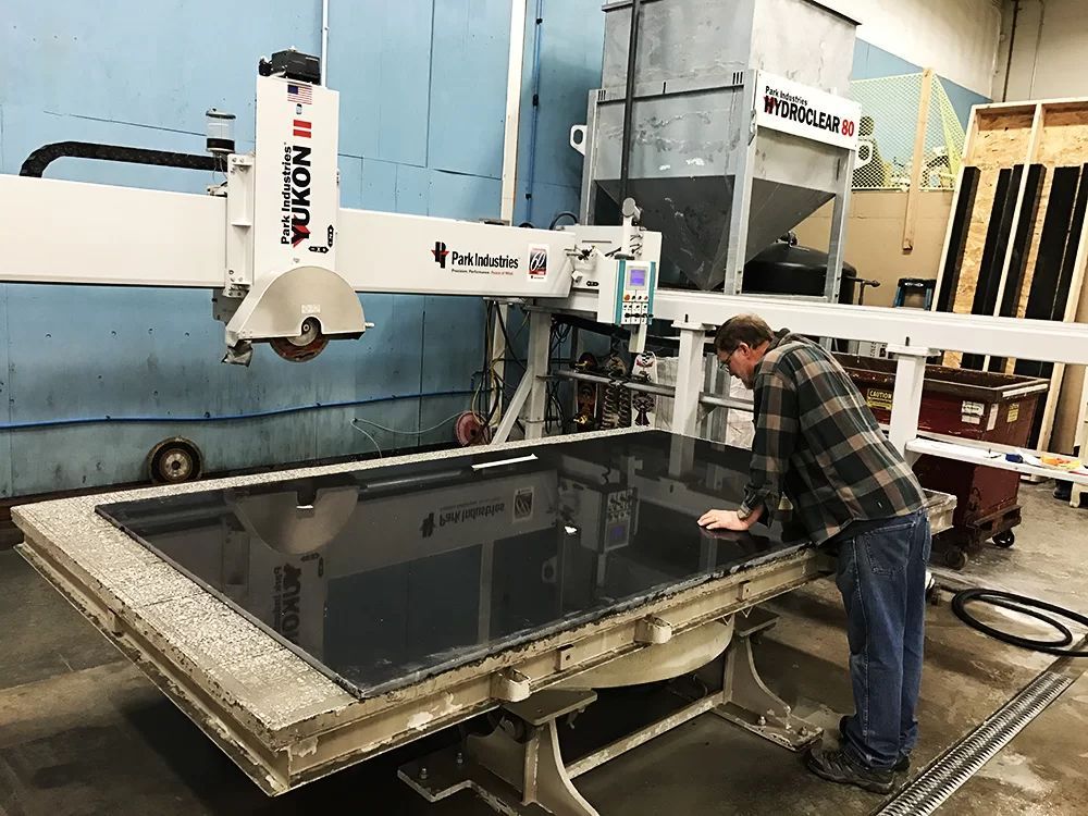 Worker operating a large industrial cutting machine in a workshop, carving a dark slab on a worktable.